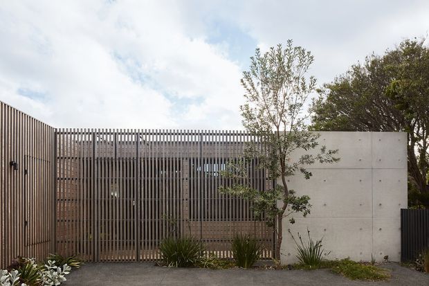 Contemporary residential facade viewed from outside, composed of vertical timber screening, a large timber slatted gate or privacy screen, and a smooth exposed concrete wall. Sparse landscaping and mature trees soften the restrained geometric frontage.