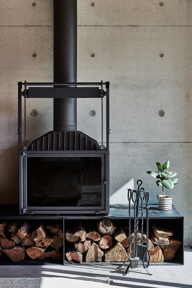 Close-up interior view of a black metal wood-burning fireplace set against a smooth exposed concrete wall with visible form-tie marks. The fireplace includes an open firebox, flue, raised frame, integrated lower timber storage filled with firewood, and a set of black fireplace tools. A small potted plant sits on the adjacent ledge, with strong sunlight casting shadows across the floor.