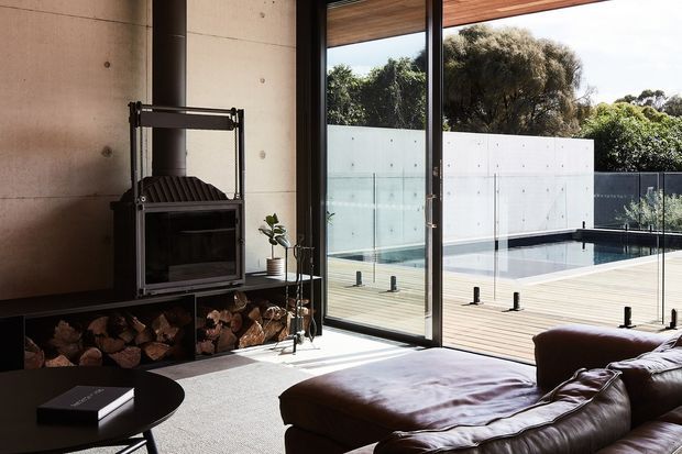 A contemporary living room with a substantial dark leather sectional sofa, low round coffee table, and a freestanding wood-burning fireplace set against a raw concrete wall. Full-height black-framed glazing opens the room visually to a timber deck and swimming pool, with a rendered privacy wall and mature planting beyond. The interior has a warm, restrained palette and strong indoor-outdoor connection.