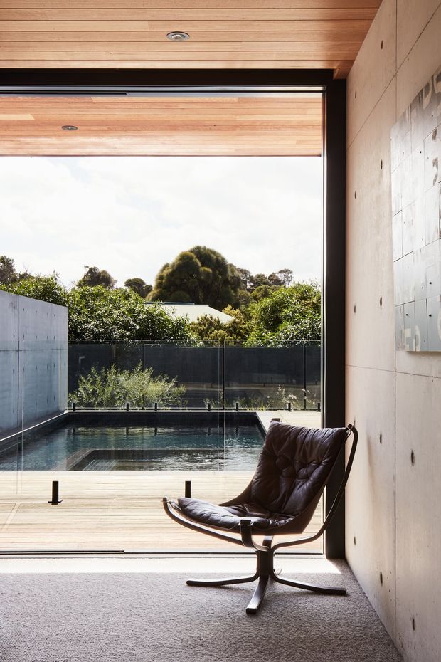 A refined indoor-outdoor living space viewed from inside through a large open glazed opening. The room has a warm timber-lined ceiling, smooth concrete walls, dark-framed glazing, and a carpeted floor. A leather sling lounge chair sits beside the opening, overlooking a timber deck, glass balustrade, and compact rectangular swimming pool surrounded by landscaped greenery. The image has a calm, modern architectural character with strong indoor-outdoor connection.