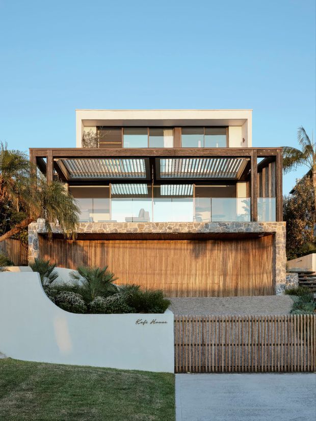 Front exterior view of a contemporary multi-storey residence with a strong indoor-outdoor connection. The facade combines extensive floor-to-ceiling glazing, a deep timber-framed pergola or sunshade, warm vertical timber cladding, and a rough natural-stone balcony wall. A gated driveway, landscaped planting, curved rendered boundary wall, and palm trees complete the street-facing composition.