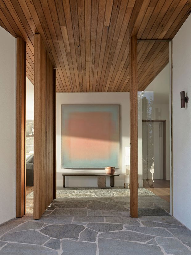 A refined residential entry hall viewed from the threshold, with irregular grey stone flooring, warm timber-clad ceiling, vertical timber door and wall elements, white plaster walls, and full-height glazing. The hall opens toward a softly lit living area featuring a low console table and abstract artwork. The design combines natural materials, strong vertical framing, and a calm, minimalist atmosphere.