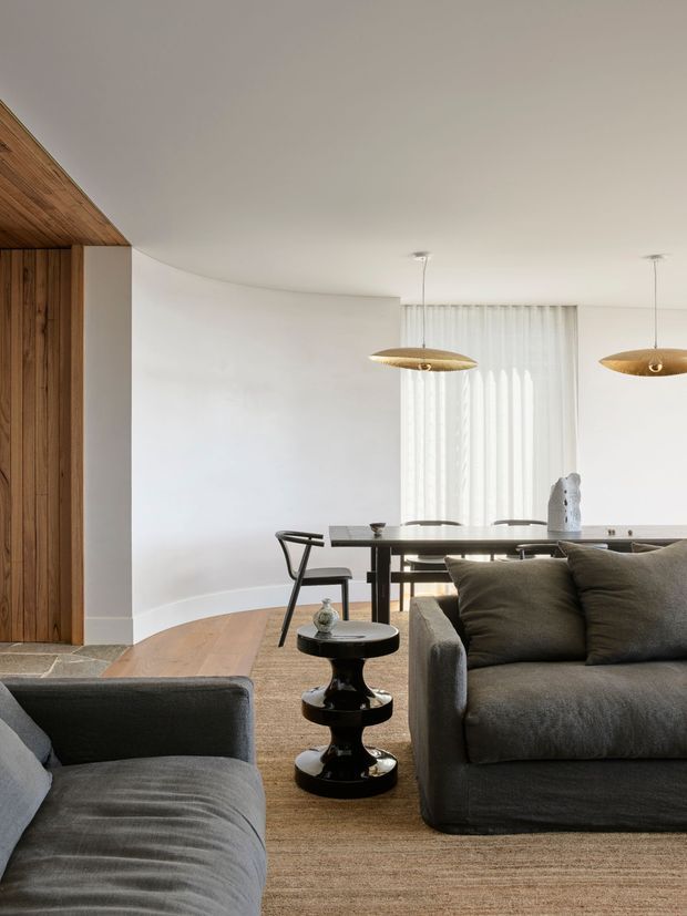 A refined open-plan living and dining area with a neutral palette. The room features a deep grey sofa and armchair, a black sculptural side table, a timber-lined built-in wall and ceiling detail, a long dark dining table, woven neutral carpeting, sheer curtains, and two organic pendant lights. A curved white wall and light timber floor create a soft, contemporary architectural backdrop.