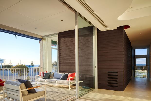 A bright contemporary indoor-outdoor living area opening onto a balcony or terrace. The space features floor-to-ceiling sliding glass doors, pale timber flooring, a white ceiling with recessed lighting and linear air vents, and a dark horizontal timber-clad built-in volume separating the living area from a hallway. Neutral outdoor-style seating with red accent cushions surrounds a low table, while a balustrade and distant urban/coastal view are visible beyond the glazing.