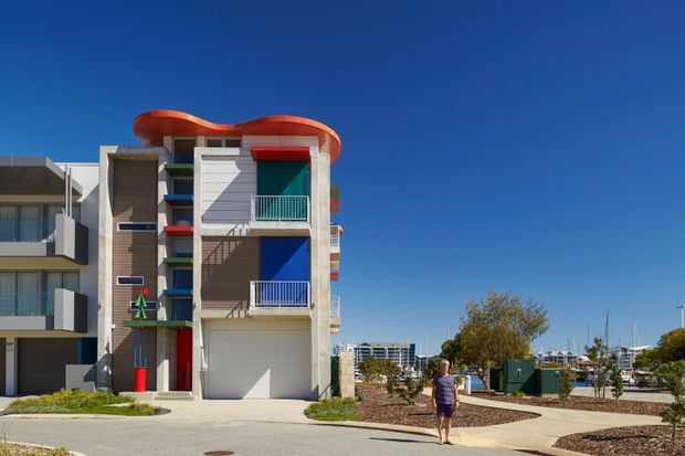Exterior view of a colourful multi-storey residential building beside a marina or waterfront setting. The facade features strong geometric forms, projecting balconies, bright red and blue accents, textured vertical cladding, white rendered walls, and a distinctive curved orange roof canopy. Adjacent apartment buildings, landscaped verges, marina infrastructure and boats are visible in the surroundings.