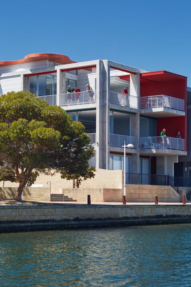 Exterior view of a contemporary multi-storey waterfront residential building with exposed concrete columns and balconies, large glazed openings, white and vivid red facade sections, and a curved roof element. The building fronts a canal or waterway with a stone retaining wall and landscaped tree in the foreground.