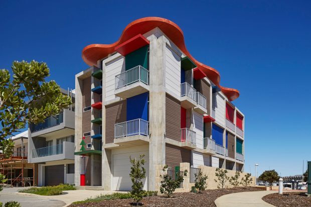 Exterior view of a contemporary multi-storey residential apartment building with an expressive undulating red roofline, exposed concrete structural elements, horizontal timber-toned cladding, bright blue and red wall accents, and multiple projecting balconies with metal balustrades. The building is set in a landscaped residential development under a clear blue sky.