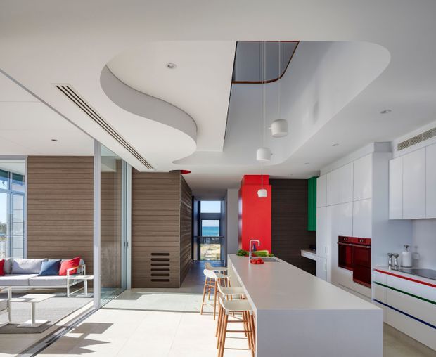 Bright contemporary open-plan kitchen and living area with a large white island, integrated cabinetry, timber wall panelling, bold red and green accent panels, and sculptural curved ceiling forms. Full-height glazing connects the living space to an outdoor view, while bar stools line the island.