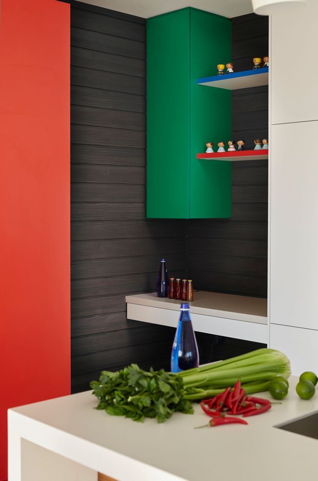 A bold, contemporary kitchen interior featuring a white island bench in the foreground, vivid red and green cabinetry, dark horizontally textured wall panelling, and white built-in cabinetry. Open display shelves hold small decorative figurines and bottles, while fresh vegetables and blue glass bottles sit on the island. The composition has a strong, playful colour-blocked design with clean lines and integrated joinery.