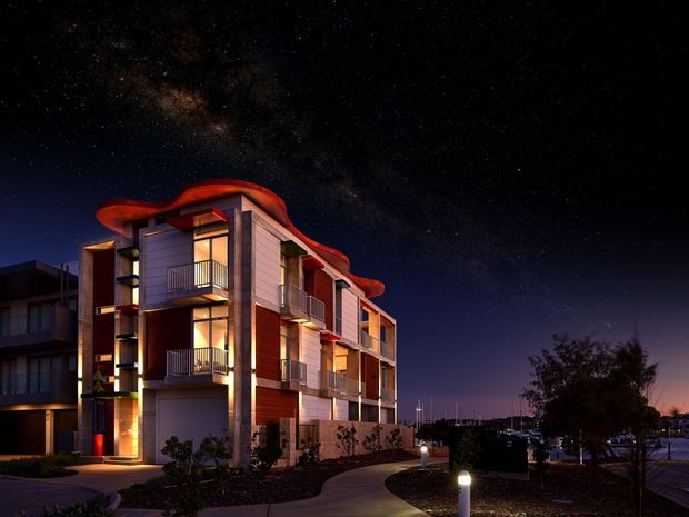 Night-time exterior view of a contemporary multi-storey residential building with illuminated balconies, extensive glazing, projecting curved roof forms, and warm architectural lighting. A landscaped pedestrian path and marina-like waterfront setting are visible beside the building under a star-filled sky.