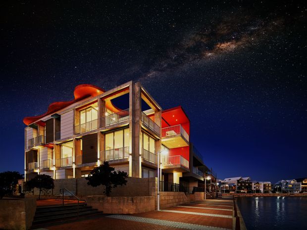 Night-time exterior view of a contemporary multi-storey residential apartment building beside a waterfront promenade. The building features strong geometric forms, projecting balconies, extensive glazing, warm illuminated interiors, concrete structural elements, rendered and panelled facades, and vivid red accent soffits. A paved pedestrian edge and water are visible in the foreground, with other residential buildings extending into the background under a star-filled sky.