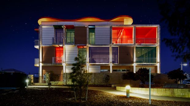 Night-time exterior view of a contemporary multi-storey residential building with a strongly sculptural, undulating roofline. The facade combines horizontal light-coloured cladding, darker vertical sections, extensive glazing, and multiple balconies, with vivid warm interior lighting visible through the windows. Landscaped planting, low retaining walls, and illuminated paths occupy the foreground.
