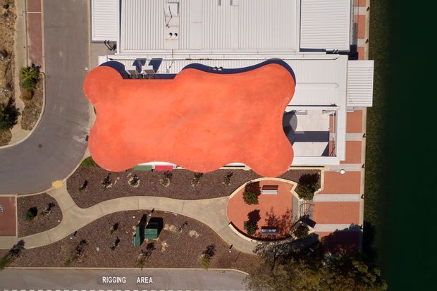 Aerial overhead view of a large building complex beside dark water, dominated by an irregular bright orange-red roof or canopy. The surrounding site includes curved pedestrian paths, landscaped planting beds, paved courtyards, road access, and a clearly marked rigging area. White and grey roof structures and red-and-beige paving are visible around the central feature.
