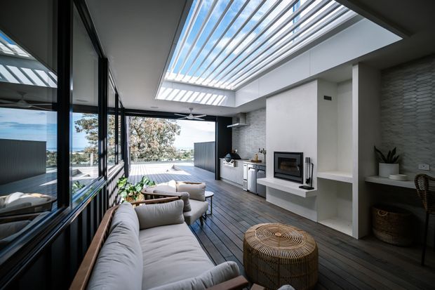 Contemporary open-plan living room with a large glazed wall, expansive operable roof louvre/skylight, built-in fireplace, and compact kitchen. A timber deck-style floor extends toward an outdoor terrace, with neutral upholstered seating, woven furniture, and views beyond the glazing.