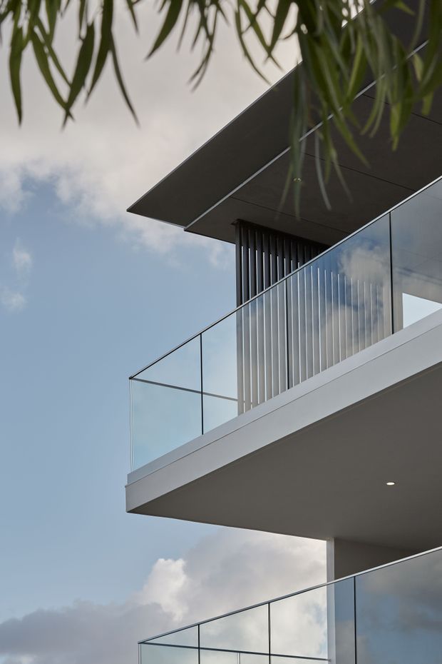 Photograph looking upward at a modern multi-level building exterior, featuring projecting flat roof slabs, crisp white and dark grey planes, extensive clear glass balustrades and glazing, and vertical metal screening. Tree foliage frames the upper edge against a partly cloudy sky.