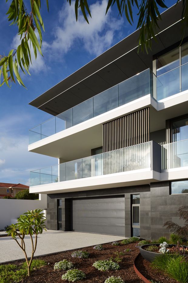 Street-level exterior view of a contemporary multi-storey residence with broad cantilevered floor slabs, extensive floor-to-ceiling glazing, clear glass balustrades, dark grey masonry cladding, white rendered surfaces, and vertical timber-look screening. A garage occupies the ground floor, with landscaped planting and a paved driveway in front. The building is photographed beneath a bright blue sky and framed by overhanging foliage.