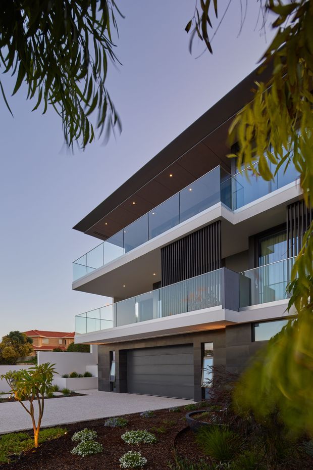A contemporary multi-storey residential facade viewed from the landscaped front yard. The house features strong horizontal floor plates, dark grey cladding and garage doors, white projecting slabs, extensive clear glass balustrades, large glazed openings, vertical screening, and a broad flat roof canopy. Low-maintenance planting and a pale paved driveway occupy the foreground, with neighbouring suburban homes visible in the background.