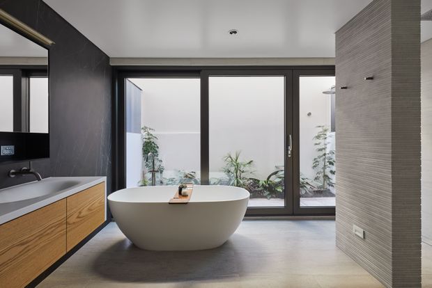 A contemporary bathroom with a freestanding white oval bathtub positioned in front of full-height dark-framed glazing, overlooking a planted courtyard. The room features a floating timber vanity with a pale benchtop, dark textured wall finishes, a grey tiled feature wall, recessed ceiling lighting, and a calm neutral palette.
