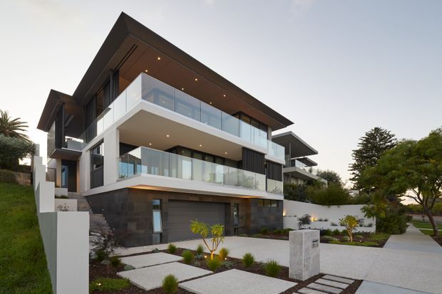 Contemporary multi-level residential house photographed from the street, featuring strong cantilevered white volumes, dark stone or masonry lower-level cladding, extensive frameless glass balustrades, dark vertical window screens, and broad roof overhangs with recessed downlights. The landscaped front approach includes pale concrete stepping slabs, low planting, a driveway or paved forecourt, and a visible garage entrance.