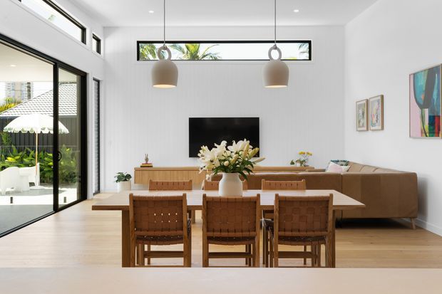 Bright open-plan dining and living area with a timber dining table and woven leather dining chairs in the foreground. A low timber sideboard, wall-mounted television, beige sectional sofa, pendant lights, large sliding glass doors, clerestory windows, and contemporary artwork complete the light-filled interior. Tropical greenery and a neighbouring roof are visible outside.