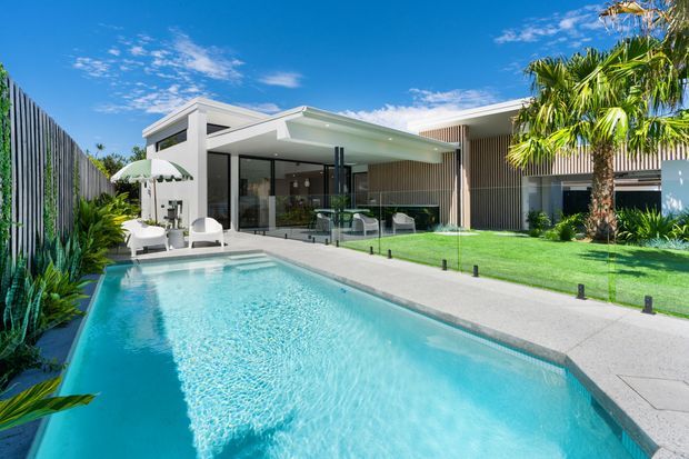 A bright contemporary single-storey residence with a long swimming pool in the foreground, landscaped lawn and palms, a covered outdoor entertaining area, and extensive glazed openings. The house combines crisp white rendered forms with dark vertical timber-like screening and a flat roofline.