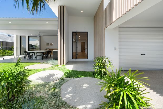 Bright contemporary residential facade featuring a recessed entry, white rendered walls, vertical timber-look screening, a large white sectional garage door, and extensive glazing opening to a covered outdoor dining area. A tropical landscaped courtyard with curved concrete stepping pads and lush planting leads to the entrance.