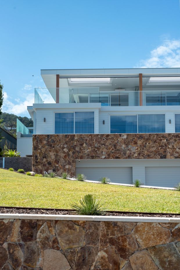 Exterior view of a contemporary multi-level residence on a sloped landscaped site. The home has crisp white horizontal cladding, extensive blue-tinted glazing, a flat roof with broad eaves and roof skylights, glass balustrades, warm timber-toned vertical supports, and prominent rough natural-stone retaining walls. A double garage is integrated into the lower level.