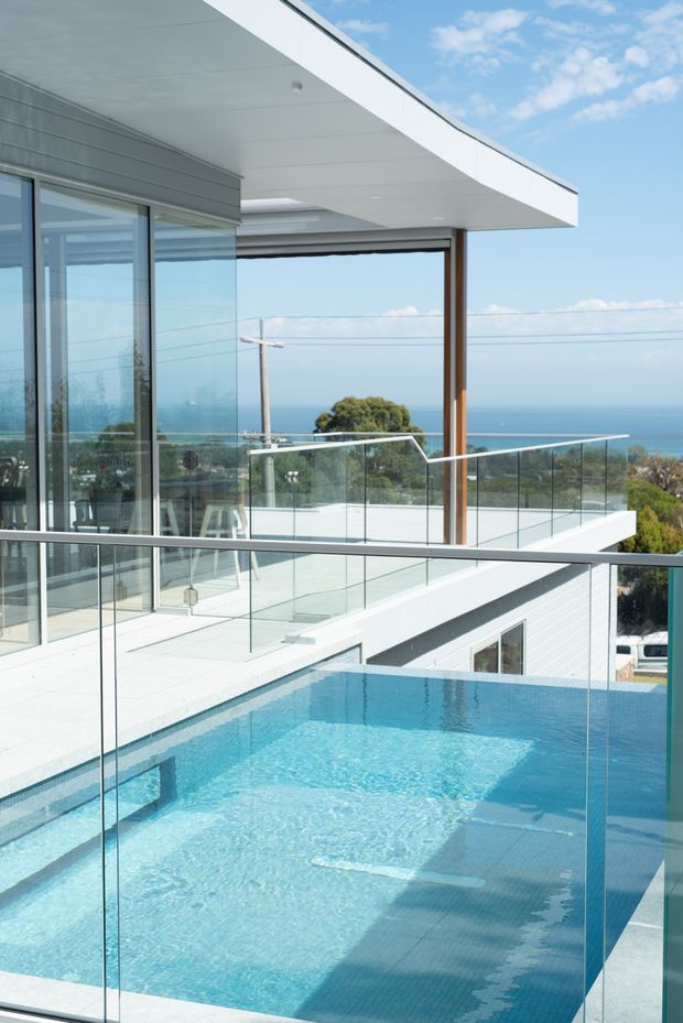 A bright contemporary coastal house exterior with a rooftop or elevated swimming pool in the foreground, surrounded by frameless glass balustrades. The building features crisp white horizontal forms, extensive floor-to-ceiling glazing, pale horizontal cladding, and a curved projecting roof canopy supported by a timber-toned vertical post. An ocean view is visible beyond the terrace, with a modern outdoor living area behind the glass doors.