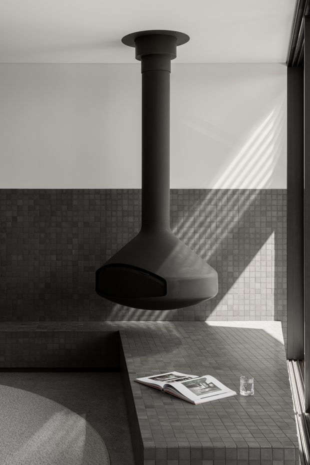 A moody, monochromatic interior featuring a suspended black cylindrical fireplace with a broad sculptural hood, set against dark small-format tiled walls and a matching tiled raised platform or hearth. Strong sunlight casts diagonal shadows through the window across the wall and surfaces. A low dark sofa or upholstered seating area is partially visible in the foreground, with an open magazine and glass on the tiled platform.