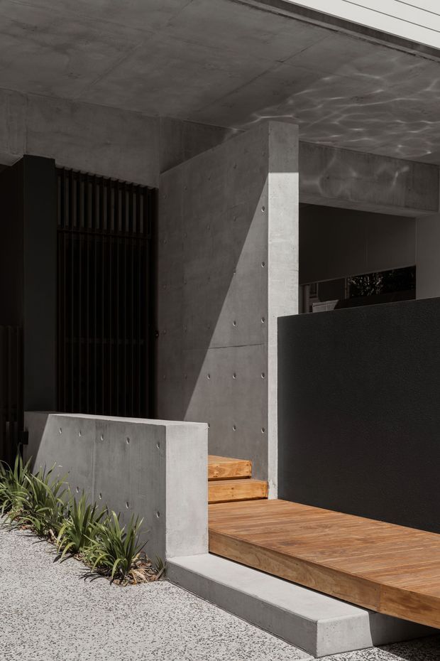 A close exterior view of a contemporary entryway featuring raw concrete walls, ceiling, columns and low retaining walls with visible form-tie marks. Warm timber decking and entry steps contrast with a dark metal security gate and dark privacy screen. Low architectural planting borders a light aggregate concrete path, with strong sunlight creating angular shadows across the facade.