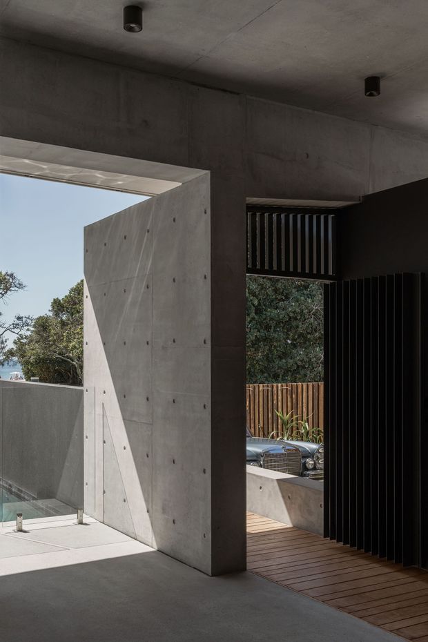 A minimalist architectural entry or passage framed by exposed grey concrete walls and ceiling, with a large open portal, black vertical slatted screen, timber decking, and glimpses of a planted exterior and pool beyond. Strong sunlight creates geometric shadows across the concrete surfaces.