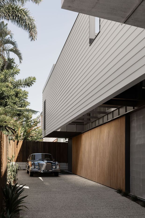 A contemporary residential exterior viewed from a driveway or carport area. The building features a strongly cantilevered upper volume clad in light horizontal weatherboards, a dark exposed soffit and structural frame, a tall warm timber garage door or wall, and a grey masonry side wall. Tropical planting and a timber boundary fence frame the narrow driveway, with a vintage car parked at the end.