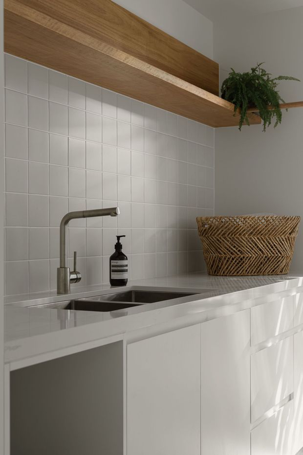 Close-up view of a refined contemporary kitchen or kitchenette, featuring white flat-panel cabinetry, a white tiled splashback, a pale stone benchtop, and a stainless-steel undermount double sink with a brushed metal tap. A warm timber overhead shelf adds contrast, with a hanging fern and woven basket providing natural texture.