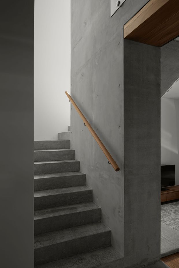 A minimalist interior stairway with exposed grey concrete steps and walls, a simple timber handrail, and a partial view into an adjacent living area with timber cabinetry and grey flooring. The stair enclosure has a strong sculptural, monolithic appearance with clean geometric lines.