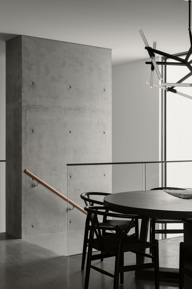 A minimalist contemporary dining area with a round black dining table and black timber chairs in the foreground. The space features a raw concrete partition wall with visible formwork joints and circular tie holes, a polished grey concrete floor, a glass balustrade, and a sculptural black-and-white linear pendant light. A timber handrail leads toward a stairway, creating an open-plan, double-level interior.