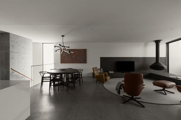 Open-plan contemporary living and dining area with a polished concrete floor, white walls and ceiling, a dark stone or textured feature wall, and extensive glazing. The space includes a round dining table with black chairs, a sculptural pendant light, a television, mid-century-style lounge chairs, a large circular rug, and a suspended black fireplace. A concrete stair wall and timber handrail are visible at left.