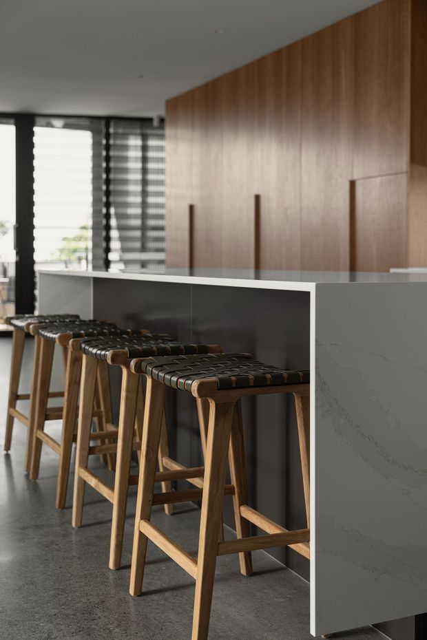 A contemporary kitchen or entertaining area featuring a long island with a pale stone waterfall end and dark charcoal side panels. Several timber bar stools with woven dark leather or strap seats line the island. Full-height warm timber cabinetry forms a clean feature wall, while large windows with layered blinds bring in filtered daylight. The floor appears to be smooth grey concrete or polished concrete, creating a refined, understated interior.