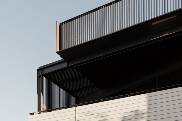 Close-up exterior architectural view of a contemporary multi-level building, showing dark metal framing, projecting balcony or roof structures, vertical metal balustrades and screens, and pale horizontal cladding. The composition emphasizes strong linear geometry and layered architectural volumes against a clear sky.