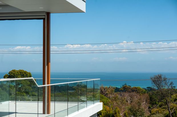 Exterior architectural view of a contemporary coastal residence, focused on a projecting balcony or terrace with frameless glass balustrades, white structural surfaces, timber-toned vertical framing, and expansive glazing. The property overlooks a blue ocean and coastal vegetation, with overhead utility lines crossing the distant view.