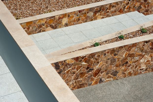 Close-up architectural detail of a stepped outdoor hardscape featuring pale square paving tiles, warm irregular natural-stone retaining walls, gravel beds, and narrow planted strips. A dark smooth wall or parapet borders the left side. The image emphasizes geometric layering, contrasting textures, and contemporary landscape construction.