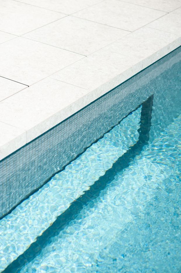 Close-up detail of a swimming pool edge, showing pale large-format paving around the perimeter, a light blue small-format tiled pool wall, and clear turquoise water with strong sunlight reflections. The image focuses on pool construction finishes rather than a broader room or landscape.