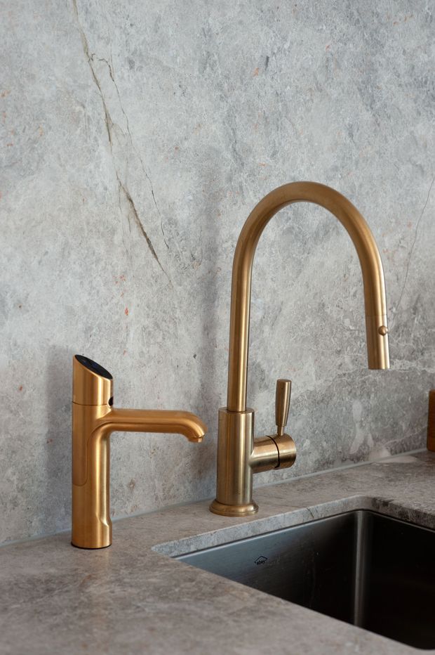 Close-up product-style view of a contemporary kitchen sink area, showing two warm bronze/gold-toned faucets mounted on a grey veined stone benchtop beside a deep dark undermount sink. The wall behind is clad in matching grey stone with natural veining.