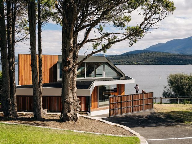 Exterior view of a contemporary multi-level house positioned beside a lake, with a steeply pitched glazed roof section, extensive timber cladding, dark metal-framed glazing, and a fenced driveway or terrace. Mature trees partially screen the building, while a mountainous shoreline is visible across the water.