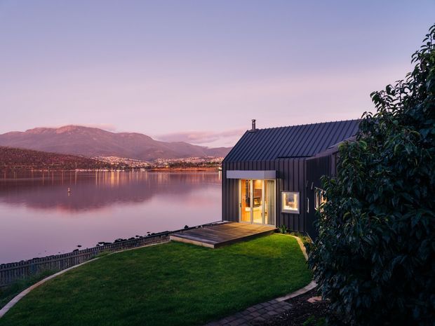 Twilight exterior view of a compact dark-clad cabin or house positioned beside a calm lake, with a pitched corrugated roof, illuminated glazing, timber deck, lawn, and a fenced waterfront edge. Mountains and reflected city lights form the background, while mature planting partially screens the building.