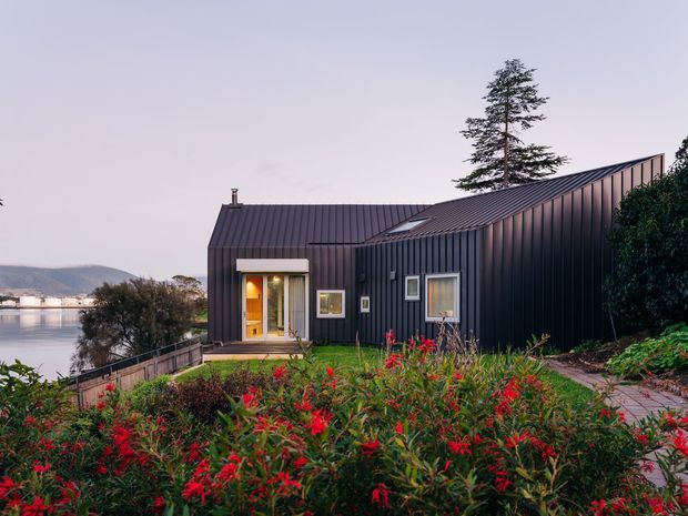 Exterior view of a compact contemporary house clad in dark vertical metal panels, with a sharply pitched standing-seam metal roof, warm-lit glazed entry, and small windows. The home sits on a landscaped sloping site beside a body of water, with flowering planting in the foreground and distant hills across the water.