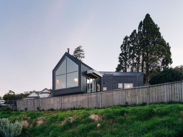 Exterior view of a contemporary single-storey house on a sloped suburban site. The dwelling has a strongly articulated dark vertical-clad form, a steep gabled glazed facade, connected low-pitched roof volumes, and extensive glazing. A timber or metal picket fence runs across the foreground, with established trees and neighbouring rooftops behind.