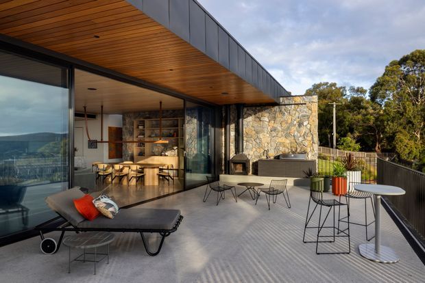 A contemporary outdoor terrace adjoining a glass-walled interior, with a broad timber-lined overhang, dark metal framing, concrete paving, and a dramatic natural-stone feature wall. The terrace contains outdoor lounge and dining furniture, bar-height tables and stools, planters, and an outdoor fireplace or built-in hearth. The interior visible through the glazing includes a dining area and timber shelving. The setting appears elevated, with distant landscape visible through the glass.