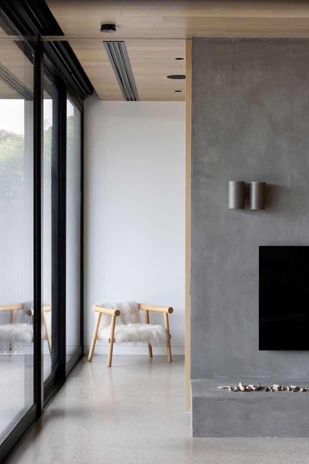 A minimalist contemporary interior corridor or living area with a polished concrete fireplace/media wall, black-framed floor-to-ceiling glazing, a pale timber ceiling, and a simple timber lounge chair with a fluffy throw. The space has clean lines, restrained neutral colours, and strong natural light.