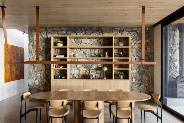 A warm, contemporary dining area with a long rounded timber dining table and multiple upholstered chairs. A full-height irregular natural-stone feature wall forms the backdrop, incorporating open timber shelving and lower cabinetry displaying books, glassware and decorative objects. The room has a timber-lined ceiling, slender timber posts, large black-framed glazing on the right, and a colourful artwork on the left.