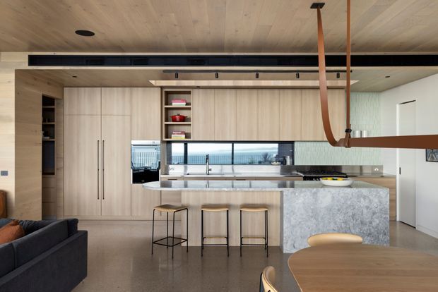 Contemporary open-plan kitchen and living area with extensive light timber cabinetry, a large grey stone island with integrated seating, polished concrete flooring, and a timber-lined ceiling. A long horizontal window provides natural light behind the benchtop, while a sculptural timber pendant hangs above the dining area. The edge of a dark sofa and a round timber dining table are visible in the foreground.